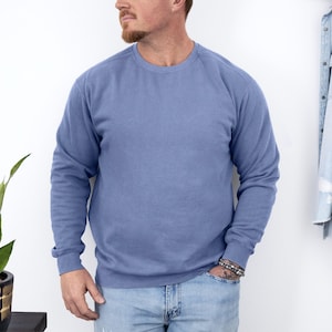 May include: A solid periwinkle blue crew neck sweatshirt. The long-sleeved top has a relaxed fit and is made of a soft material. The sweatshirt is paired with light blue jeans. A denim jacket hangs on the wall in the background.