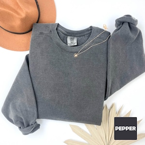 May include: A dark gray sweatshirt with a crew neck and long sleeves. The sweatshirt is made of a soft, comfortable fabric and has a vintage look. The word "COMFORT COLORS" is printed on the tag.