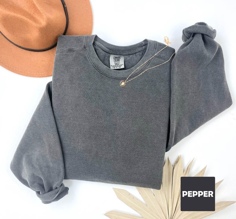 May include: A charcoal gray crewneck sweatshirt with a folded sleeve, a gold necklace, and a brown hat. The sweatshirt has a tag on the collar and the word "PEPPER" in a black box. The sweatshirt is made of a soft material.