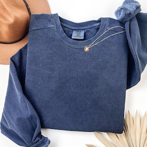 May include: A navy blue crew neck sweatshirt with the Comfort Colors brand tag. A gold necklace with a pendant is draped across the front. A brown hat is visible in the upper left corner.