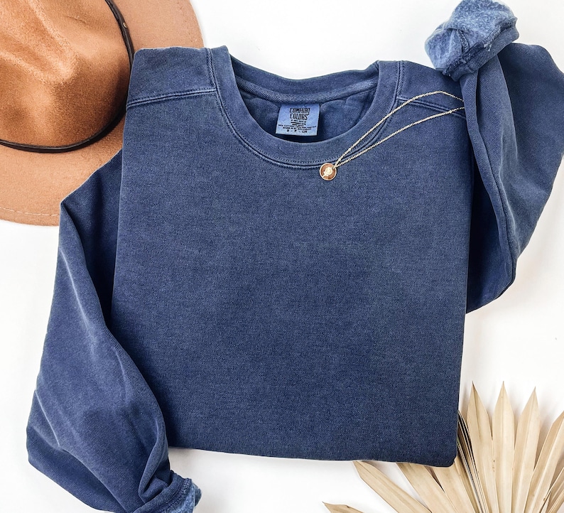 May include: A navy blue crew neck sweatshirt with the Comfort Colors brand tag. A gold necklace with a pendant is draped across the front. A brown hat is visible in the upper left corner.