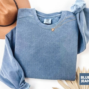May include: A blue jean-colored crewneck sweatshirt with a gold necklace. The sweatshirt has a faded, vintage look. The tag reads "Comfort Colors". A brown hat is visible in the background.