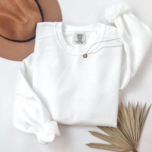 May include: A white crewneck sweatshirt with the tag "Comfort Colors" is displayed. A gold-colored necklace with a circular pendant rests on the sweatshirt. A brown hat and dried palm leaves are also in the image.