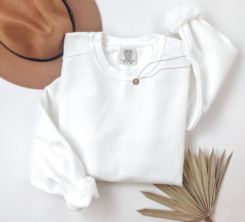 May include: A white crewneck sweatshirt with the tag "Comfort Colors" is displayed. A gold-colored necklace with a circular pendant rests on the sweatshirt. A brown hat and dried palm leaves are also in the image.