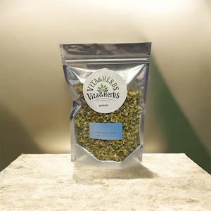 May include: A clear, sealed bag of dried chamomile flowers. The bag features a round label with "Vita & Herbs" and a blue label reading "Chamomile Flower". The bag is on a textured surface.