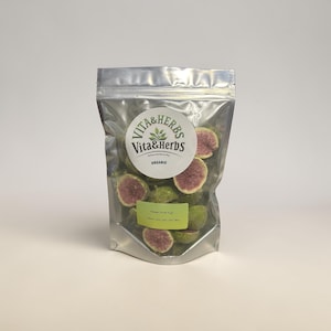 May include: A clear, metallic bag filled with freeze-dried figs. The bag has a circular label with the text "Vita & Herbs" and "Organic". The figs are green with a pink interior. A green label at the bottom of the bag reads "Freeze Dried Figs".
