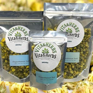 Op de afbeelding: Drie doorzichtige, verzegelde zakken met biologische chrysantenknoppen van Vita & Herbs. Elke zak heeft een wit etiket met het Vita & Herbs-logo en het woord "ORGANIC". Een blauw etiket op elke zak luidt "Chrysanthemum Buds".