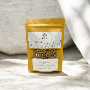 May include: A golden-yellow pouch of Vita Herbs Organic Raspberry 1000mg capsules. The clear window reveals numerous brown capsules. The label features the brand name and botanical illustrations. The pouch is sealed and stands upright on a light-colored surface.