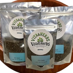 May include: Three clear, sealed bags of chia seeds, labeled "Vita & Herbs Organic," are arranged in a wooden bowl. Each bag has a blue label with "Chia Seeds" printed on it. The seeds are dark and fill the bags.