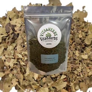 May include: A clear plastic bag filled with dried raspberry leaf, labeled "Dried Raspberry Leaf" and "Organic" by Vita & Herbs. The bag is surrounded by a pile of dried leaves, with a white background.