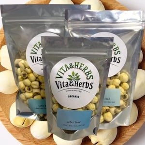 May include: Three clear, resealable bags of lotus seeds, labeled "Vita & Herbs Organic," are arranged on a wooden tray with several white lotus seed pods. The bags have a circular logo and a blue label that reads "Lotus Seed."