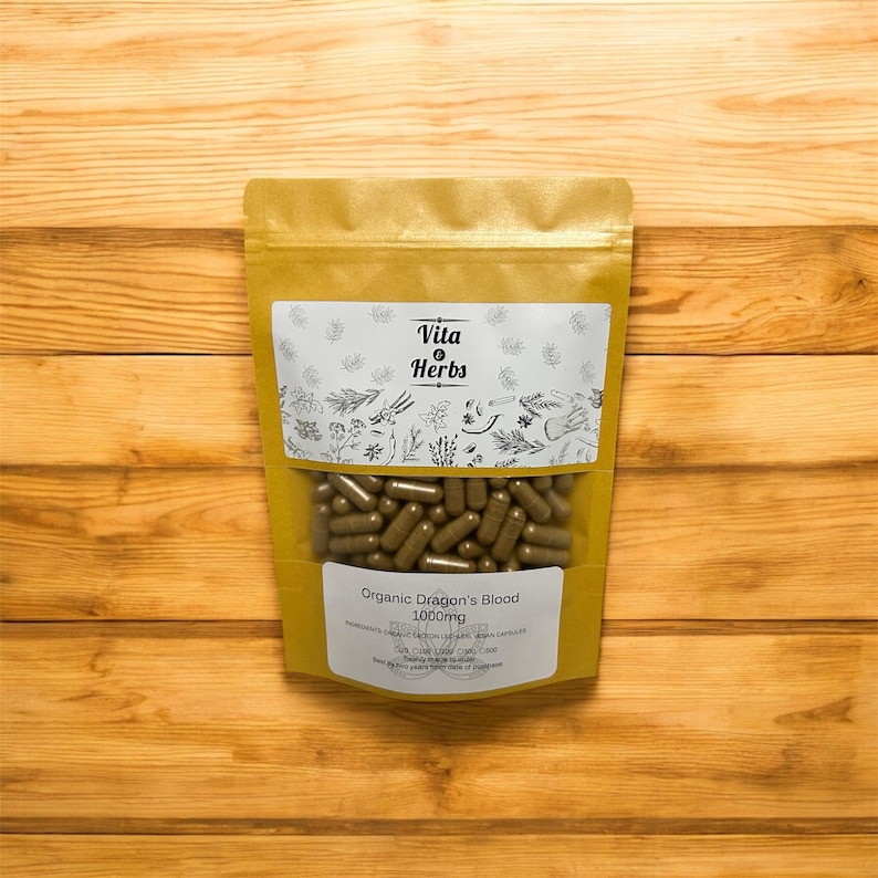 May include: A gold-colored pouch of Vita Herbs capsules, labeled "Organic Dragon's Blood 1000mg." The pouch has a clear window showing numerous brown capsules. The background is a light brown wooden surface.