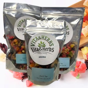 Puede incluir: Tres bolsas transparentes de té de frutas Vita & Herbs, cada una con una etiqueta redonda. Las etiquetas dicen "Vita & Herbs" y "Organic". Las bolsas están llenas de frutas secas de colores. Trozos de fruta sueltos están esparcidos.