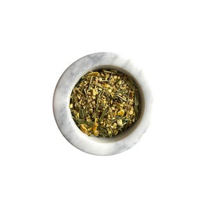 May include: A white marble pestle and mortar containing a mix of dried herbs and spices. The mixture includes green, yellow, and brown pieces. The mortar is round with a smooth, polished surface, suggesting a blend of ingredients.