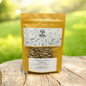 May include: A brown paper bag of Vita Herbs organic clove capsules, 1000mg. The bag has a clear window showing the capsules. The background is a blurred green, and the bag is resting on a wooden surface.