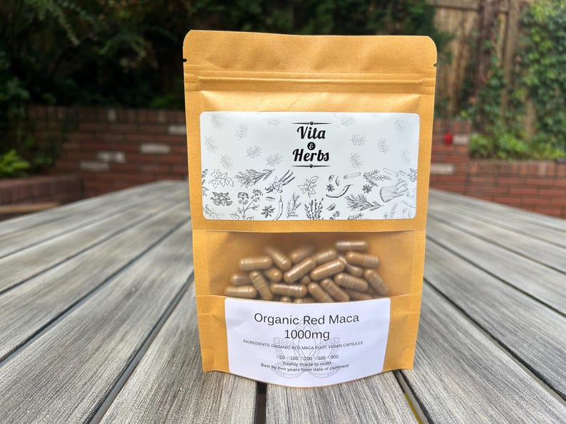 Op de afbeelding: Een bruine papieren zak met een wit label waarop "Vita Herbs" en "Organic Red Maca 1000mg" staat. De zak bevat bruine capsules.