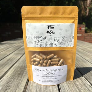 Pu&ograve; includere: Un sacchetto di carta marrone con un'etichetta bianca che dice "Vita & Herbs" e "Organic Ashwagandha 1000mg". Il sacchetto contiene capsule marroni.