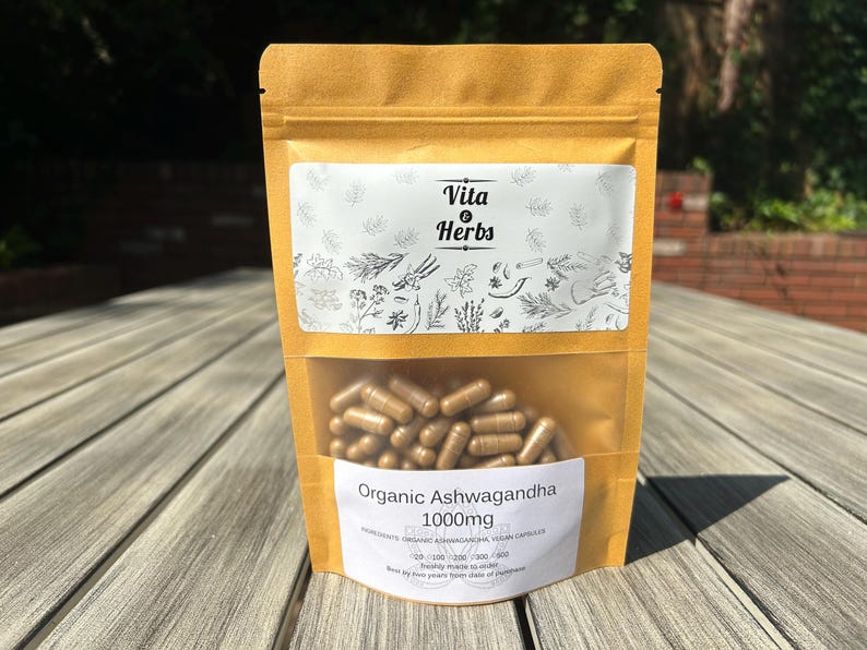 Pu&ograve; includere: Un sacchetto di carta marrone con un'etichetta bianca che dice "Vita & Herbs" e "Organic Ashwagandha 1000mg". Il sacchetto contiene capsule marroni.