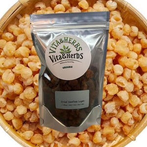 May include: A clear bag of dried seedless longan, labelled "Vita & Herbs Organic," is in a woven basket surrounded by dried longan fruit. The bag is held in a silver metal holder. The basket is made of light brown wood.