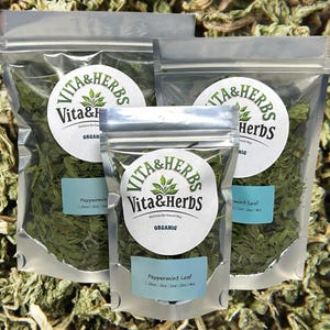 May include: Three silver resealable bags filled with dried peppermint leaves. The bags have a white label with the text "Vita & Herbs" and "Organic" printed on them. The bags are sitting on a bed of dried peppermint leaves.