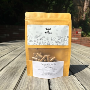 May include: A tan pouch with a clear window displaying brown capsules. The pouch has two white labels with text and botanical illustrations. The top label reads "Vita Herbs." The bottom label says "Organic Amla 1000mg."