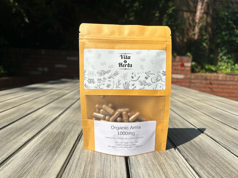 May include: A tan pouch with a clear window displaying brown capsules. The pouch has two white labels with text and botanical illustrations. The top label reads "Vita Herbs." The bottom label says "Organic Amla 1000mg."