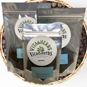 May include: Three clear plastic pouches of black peppercorns inside a woven basket. Each pouch has a round label with the text "Vita & Herbs" and "Organic". The basket is filled with peppercorns, and the background is white.