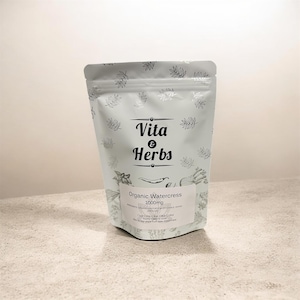 以下が含まれることがあります： 「Vita & Herbs」と「Organic Watercress 1000mg」の文字が入った水色のポーチ。ポーチには銀色の葉模様とハーブのイラストが描かれています。ポーチは明るい色の表面に立っています。