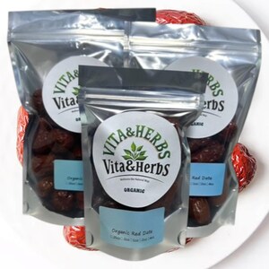 Może przedstawiać: Trzy przezroczyste torebki wypełnione organicznymi czerwonymi daktylami, każda z okrągłą etykietą z napisem "Vita & Herbs". Torebki ułożone są na białym talerzu, z kilkoma czerwonymi daktylami widocznymi wokół.