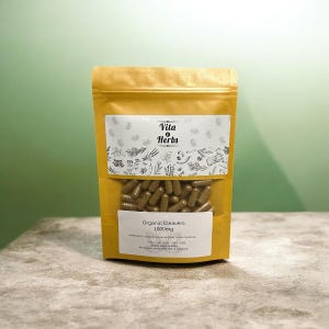May include: A gold-colored pouch of Vita Herbs Organic Cleavers capsules. The pouch has a clear window showing the capsules. The label reads "Organic Cleavers 1000mg". The background is a light green wall and a gray surface.