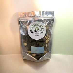 Puede incluir: Una bolsa plateada sellada llena de setas de colmenilla secas. La bolsa tiene una etiqueta redonda con el texto "Vita & Herbs" y un diseño de hojas. Una pequeña etiqueta azul dice "Wild Morels". El fondo es un degradado de rosa claro y verde.