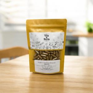 May include: A gold-colored pouch of Vita Herbs Organic Sprouted Broccoli capsules. The pouch has a clear window showing the brown capsules. The label reads "Organic Sprouted Broccoli 1000mg". The background is a wooden table and a blurred kitchen.
