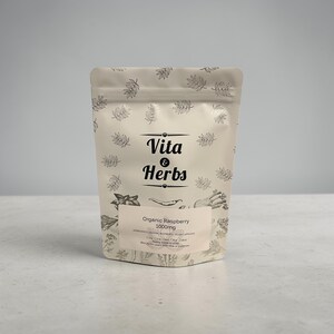 May include: A white pouch with a leaf pattern and the text "Vita & Herbs." The pouch contains "Organic Raspberry 1000mg" capsules. The background is a light gray.