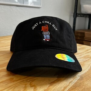 May include: Black baseball cap with an embroidered cartoon character and the text "JUST A CHILL GUY".