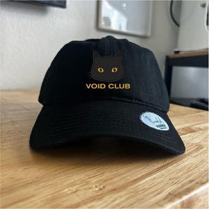 May include: A black baseball cap featuring an embroidered graphic of a black cat head with yellow eyes and the text VOID CLUB in yellow lettering. A small circular sticker is attached to the right side of the curved brim of the cap.