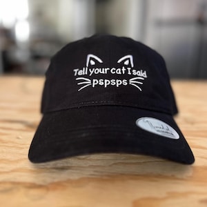Puede incluir: Gorra de béisbol negra con bordado blanco que dice "Tell your cat I said pspspps" y una cara de gato con bigotes.