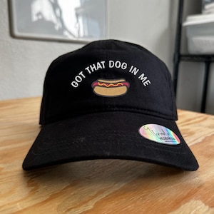 Puede incluir: Gorra de béisbol negra con texto blanco que dice "GOT THAT DOG IN ME" y un gráfico de hot dog de dibujos animados.