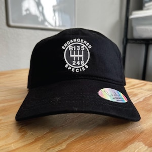 May include: Black baseball cap with the text "ENDANGERED SPECIES" and a gear shift pattern graphic in white. The cap has a curved brim and a holographic sticker on the side.