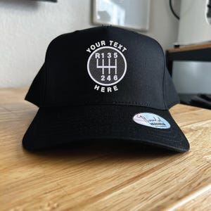 May include: Black baseball cap with a flat brim. The cap features white embroidered text and a gear shift pattern with the words "YOUR TEXT" above and "HERE" below. The gear shift pattern includes the numbers R135 and 246.