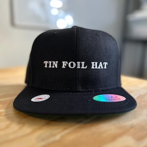 Może przedstawiać: Czarna czapka bejsbolówka z haftowanym białym napisem "TIN FOIL HAT" z przodu.