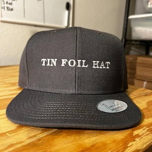 Może przedstawiać: Czarna i szara czapka z daszkiem z haftem "TIN FOIL HAT" w kolorze białym z przodu.