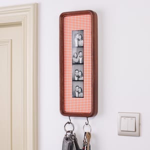 Op de afbeelding: Een rechthoekige houten wand-sleutelhouder met een verticale fotostrip van een koppel in een lijst met een oranje en wit ruitjespatroon. Twee metalen haken aan de onderkant houden verschillende metalen sleutelringen en sleutels vast.
