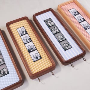 Op de afbeelding: Drie rechthoekige houten wandframes met afgeronde hoeken, elk voorzien van een verticale strook van vier zwart-wit fotohokje-afbeeldingen gecentreerd op een achtergrond van gele, blauwe of oranje gingham-stof.