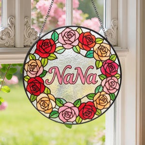 Puede incluir: Un adorno de ventana redondo de vidrieras con un marco negro. Muestra una corona de rosas rojas, rosas y amarillas que rodean la palabra "NaNa" en escritura rosa. Se adjunta una cadena para colgar.