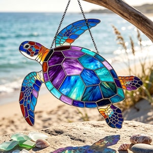 Puede incluir: Un atrapasoles de vidriera con forma de tortuga marina, compuesto por un mosaico vibrante de segmentos de vidrio morados, azules y turquesas delineados en metal oscuro. La tortuga está suspendida por una fina cadena de color negro.