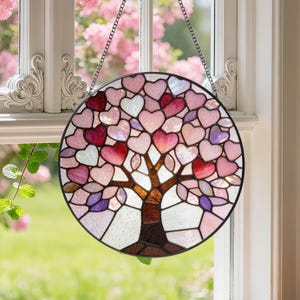 May include: Round stained glass window art featuring a tree design with heart-shaped leaves in shades of pink, red, and white. The tree trunk and branches are brown. The window hangs from a chain in front of a window.