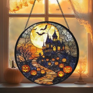 Może przedstawiać: Okrągła witrażowa dekoracja na Halloween przedstawiająca nawiedzony zamek, dynie i nietoperze pod pełnią księżyca. Grafika jest oprawiona w czarną ramę i zawieszona na łańcuchu. Dynia leży na parapecie.