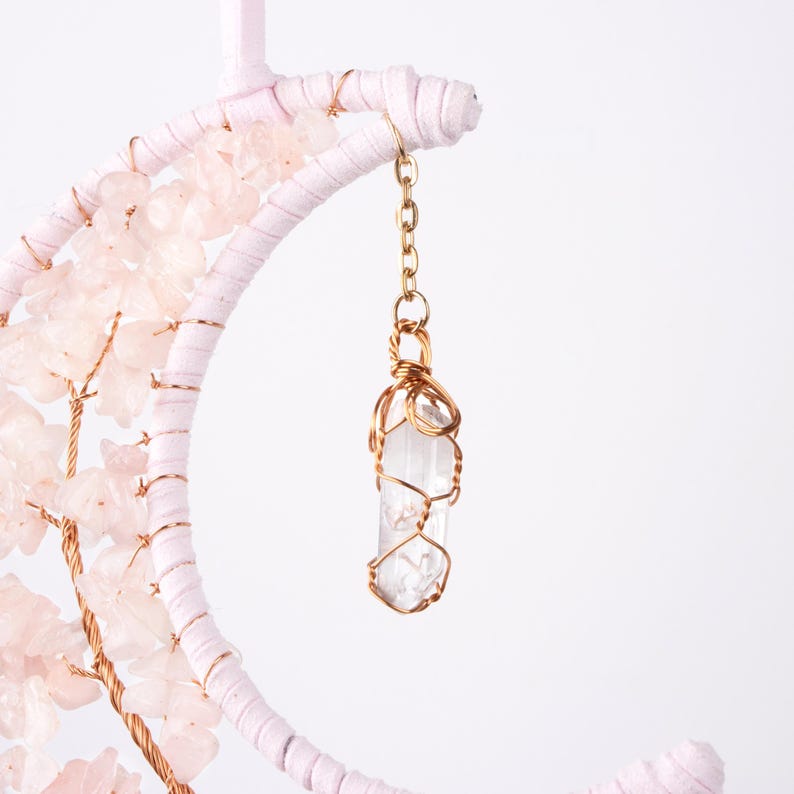 May include: A white crescent moon with pink crystals and a clear quartz crystal wrapped in gold wire hanging from it.