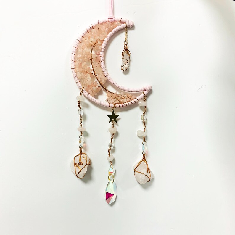 May include: A pink and gold wire-wrapped moon with crystals hanging from it. The moon is decorated with rose quartz crystals and has a gold star in the center. The crystals hanging from the moon are clear and iridescent.