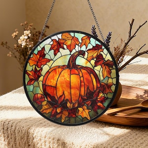 May include: A circular stained glass window depicting a pumpkin and autumn leaves in orange, red, and yellow hues. The pumpkin is central, encircled by colourful maple leaves. A black metal chain is attached for hanging.